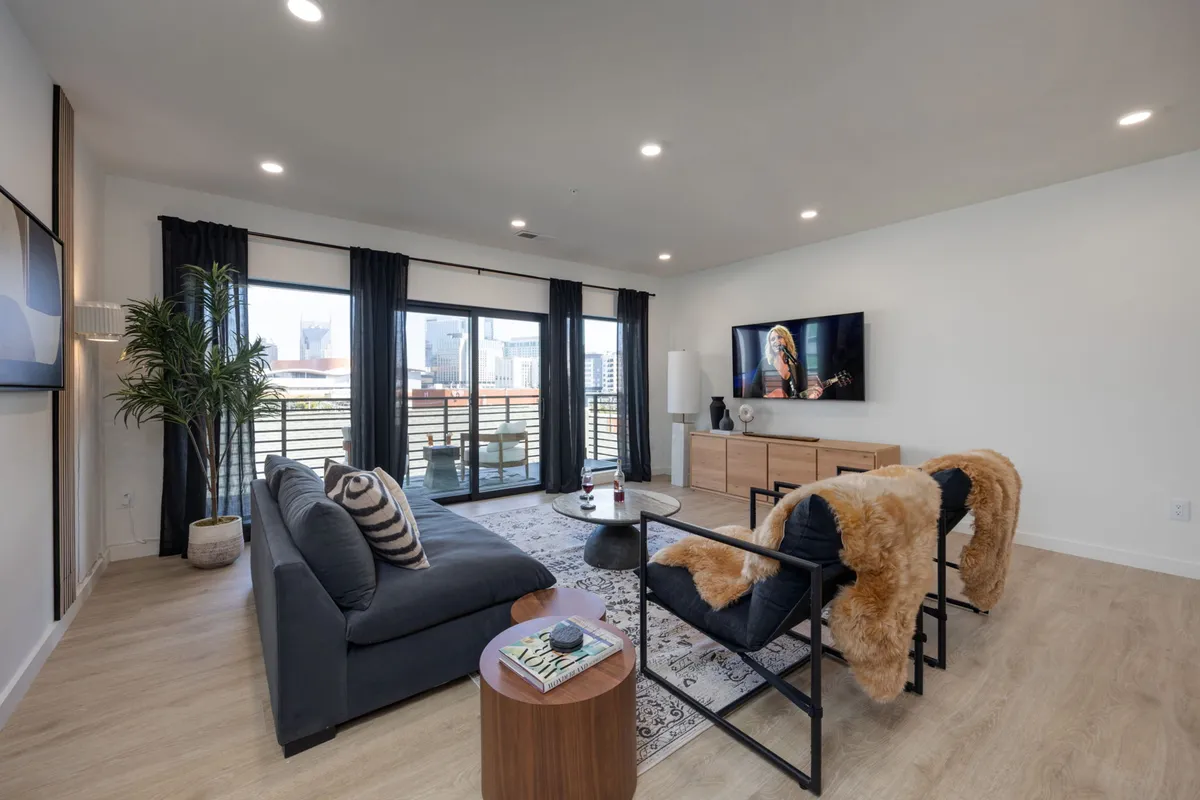 The image showcases a stylish, modern living room with large windows framing city views, a plush sofa, a sleek television, a decorative plant, and a coffee table—inviting spaces perfect for gathering, relaxing, and enjoying the Nashville vibe.