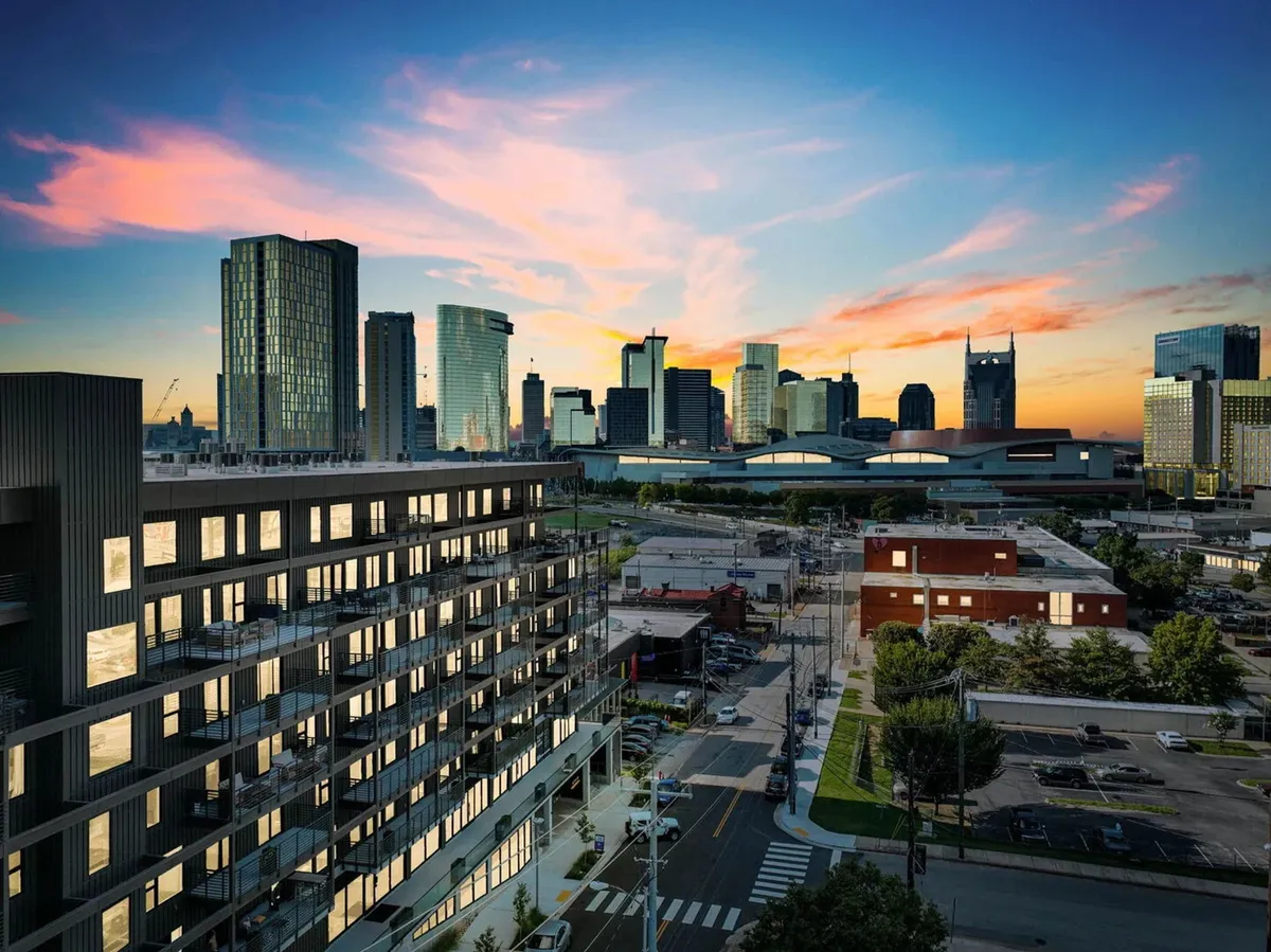 A modern apartment building with a dramatic city skyline and a colorful sunset overhead. The foreground hints at evening strolls and nearby dining, while the background radiates the inviting energy of a lively urban neighborhood.