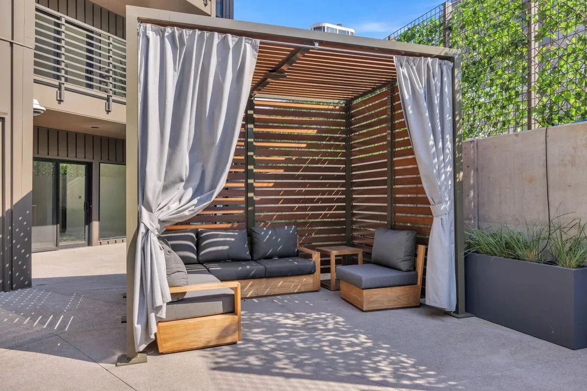 An exterior shot of a private patio with a wooden pergola, cushioned seating, curtains for privacy, and planters. The space is both cozy and inviting, ideal for enjoying morning coffee or evening conversations under the city sky.