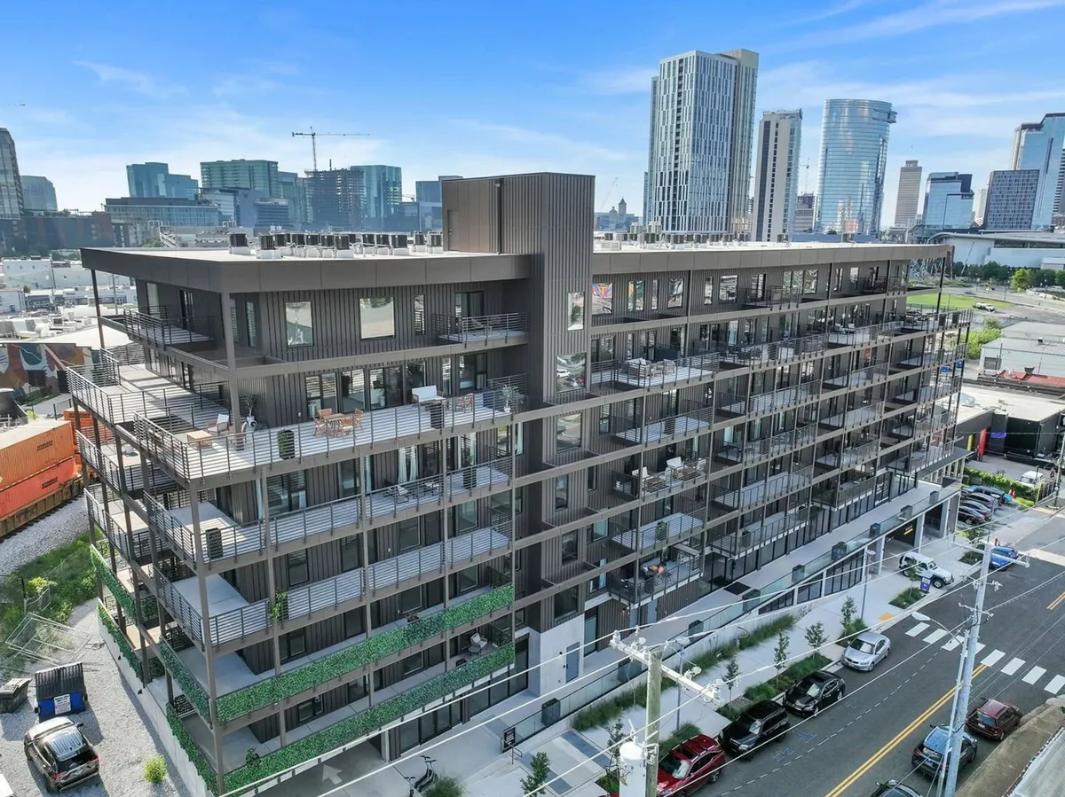 The exterior of a modern apartment building with prominent balconies and greenery, set against a lively city skyline. The pedestrian-friendly environment and visible garage entry convey security, urban vibrancy, and convenient access to parking.