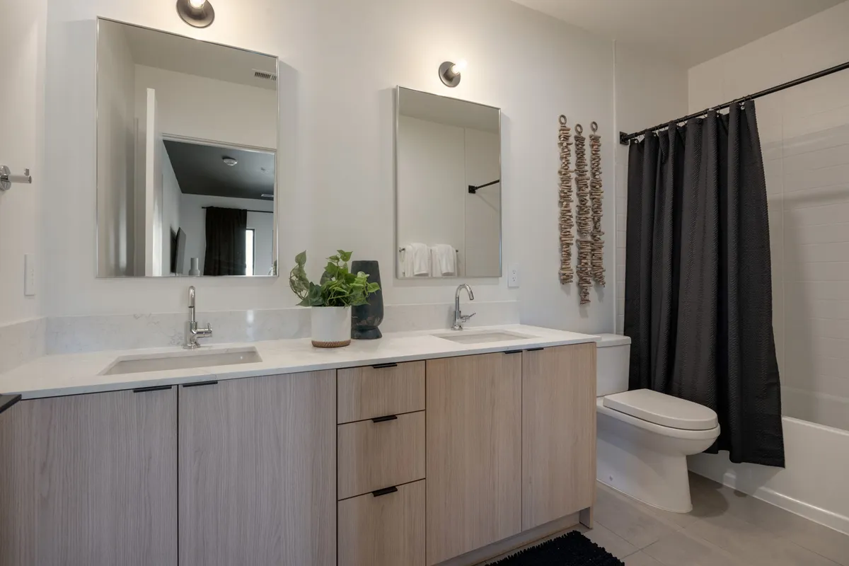 A serene, spa-inspired bathroom featuring a double sink vanity, large mirrors, potted plant, and a sleek black shower curtain. The clean lines and thoughtful details evoke a boutique hotel experience.