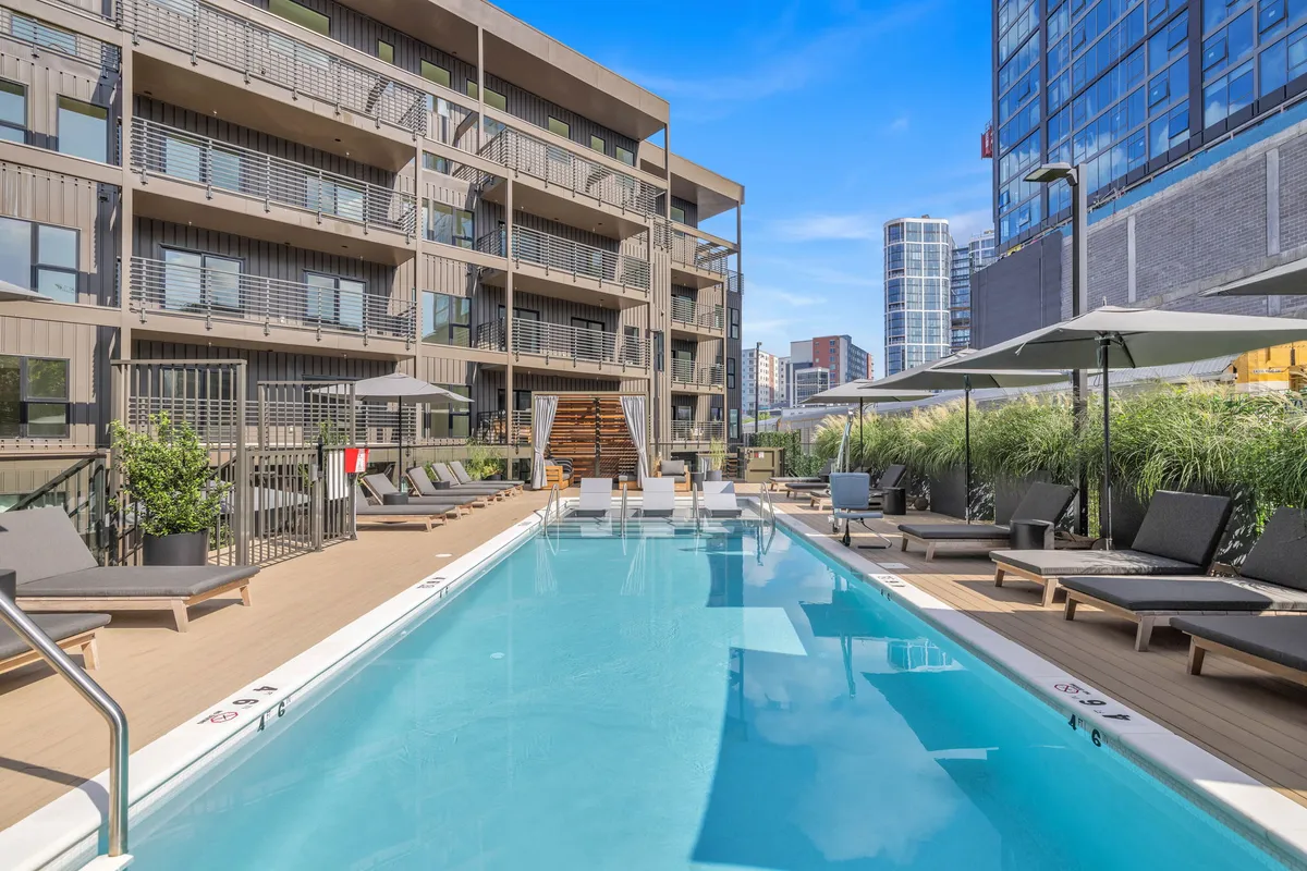 A chic, inviting swimming pool area framed by modern lounge chairs, the building’s facade, and lush landscaping. The open sky overhead and well-placed greenery create a luxurious, resort-like outdoor amenity space that encourages relaxation and social gatherings.