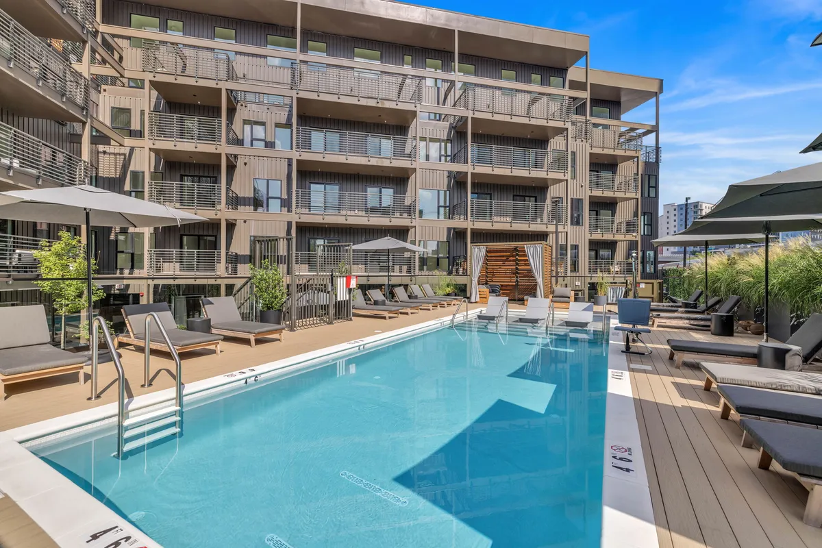 The image depicts a sun-soaked swimming pool area surrounded by lush greenery and modern building facades. Outdoor seating and social spaces suggest opportunities for relaxation and group activities within the property.