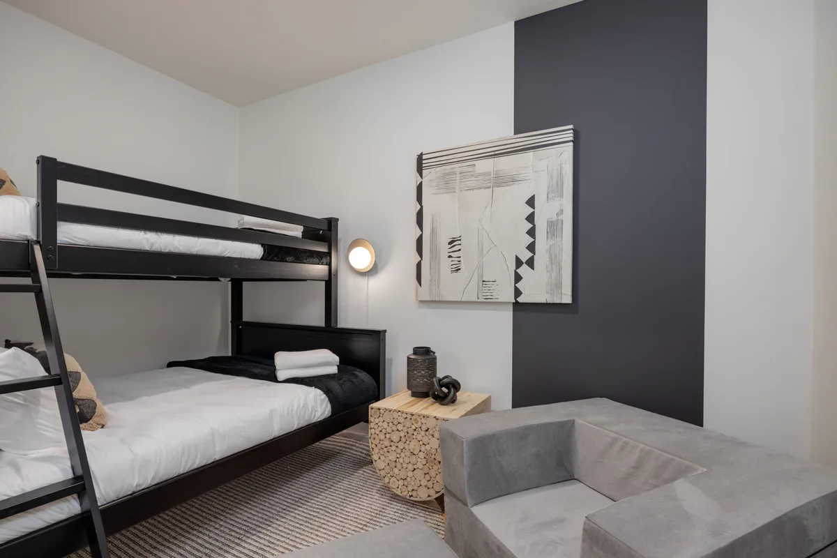 A second bedroom styled with a queen/twin bunk bed, abstract wall art, an inviting accent chair, and a wooden side table. The room blends playful elements with modern design, making it ideal for families or friends and showcasing flexible sleeping arrangements.