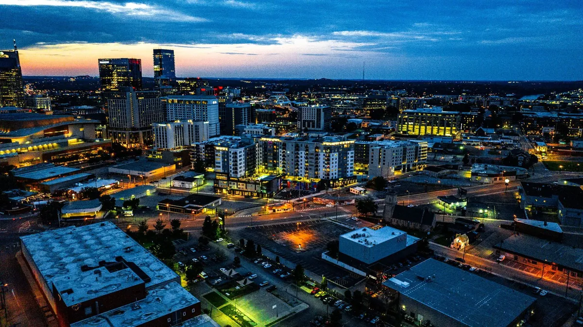 A dazzling city skyline is illuminated at dusk, with buildings, street lights, and parking lots glowing against the evening sky. The scene captures the vibrant city atmosphere that becomes the backdrop for cozy evenings or movie nights inside.