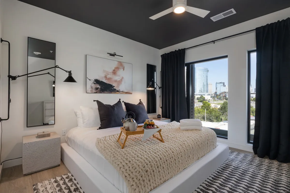 A modern, cozy bedroom featuring a plush bed with a textured blanket, large windows offering a city view, decorative wall art, and stylish lighting fixtures. The morning light filters softly into the room, enhancing the restful and inviting atmosphere.