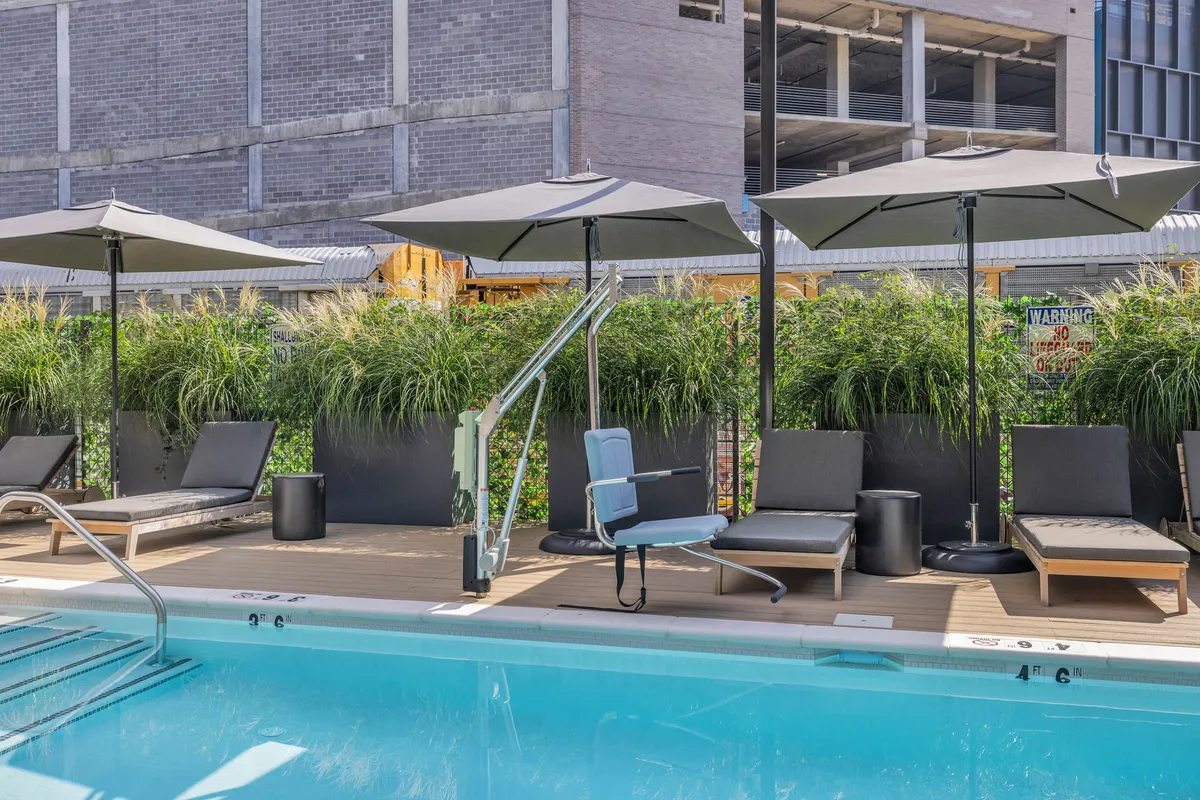 A sunlit swimming pool is surrounded by lounge chairs, umbrellas, and contemporary planters filled with decorative grass. The pool deck is spacious, offering areas for sunbathing, swimming, and socializing. The scene captures a tranquil atmosphere, suggesting both relaxation and community gathering under a blue sky.