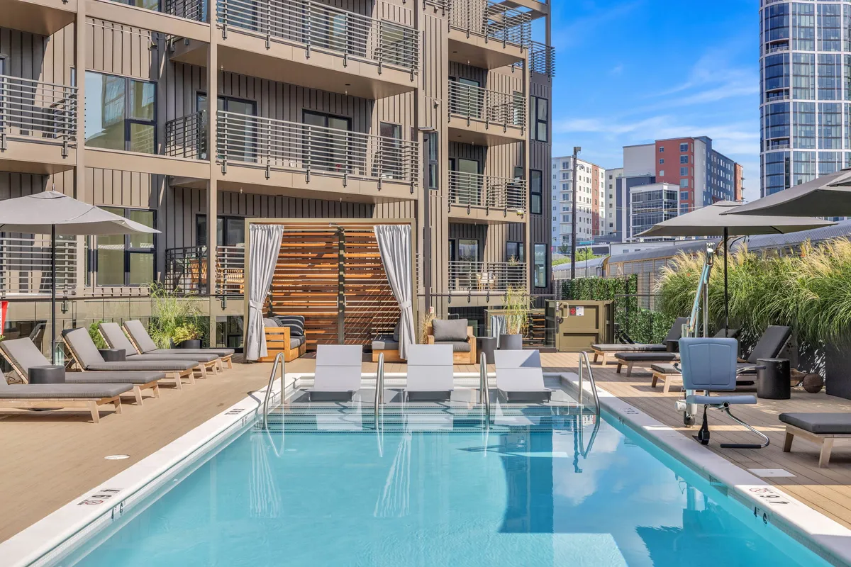 This image features a sparkling swimming pool surrounded by lounge chairs, umbrellas, and the building facade in the background. The mood is leisurely, with people swimming, sunbathing, and socializing, creating an inviting atmosphere for relaxation and community gatherings.