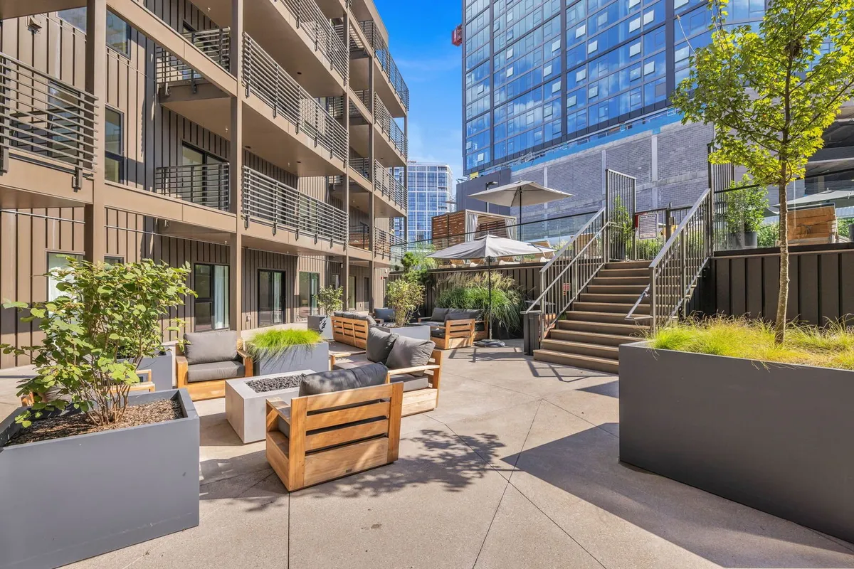 A contemporary outdoor seating area with planters, sleek furniture, and a staircase leading up to a modern building. The space is ideal for gatherings, casual conversations, or a tranquil morning coffee, surrounded by greenery and urban architecture.